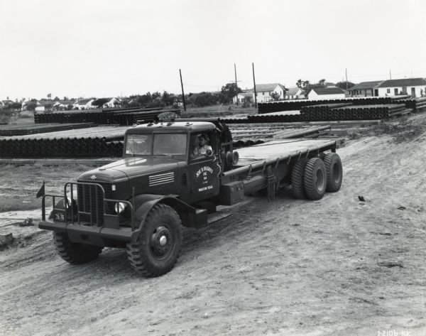 Elevated three-quarter front view of an International truck owned by Joe D. Hughes Incorporated. A man sits behind the wheel of the truck parked in a dirt lot in front of stacks of metal pipes.