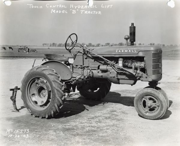 Engineering photograph of right side of a Farmall B tractor parked in a dirt lot. Cows and an additional tractor are in a field in the background. The text on the photograph reads: "Touch Control Hydraulic Lift Model 'B' Tractor."