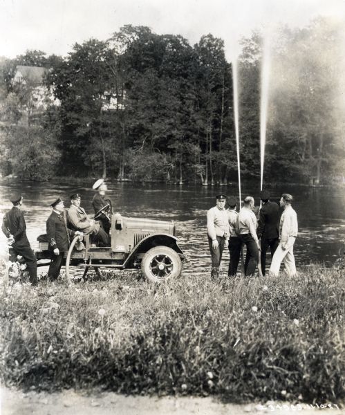 Several men are sitting and standing around an International truck with pump(?) while other men are operating fire hoses shooting water up into the air on the shore of a body of water.