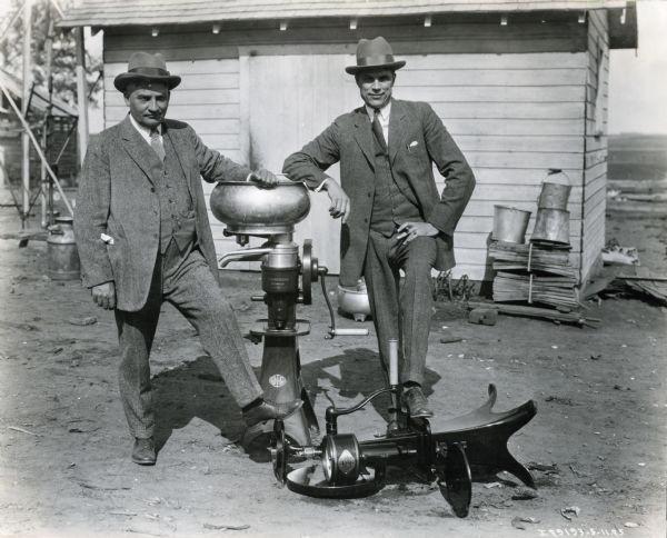 Two men are posing outdoors wearing three-piece suits. They are standing on either side of a McCormick cream separator. An additional cream separator is lying on its side on the ground, and on which both men have placed their feet. The men may be International Harvester dealers or salesmen.