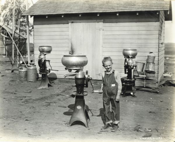 A young boy named Lowell Schmidt is turning the handle of a cream separator in front of a farm building. Two additional cream separators are on either side of the boy in the background, along with several milk pails.