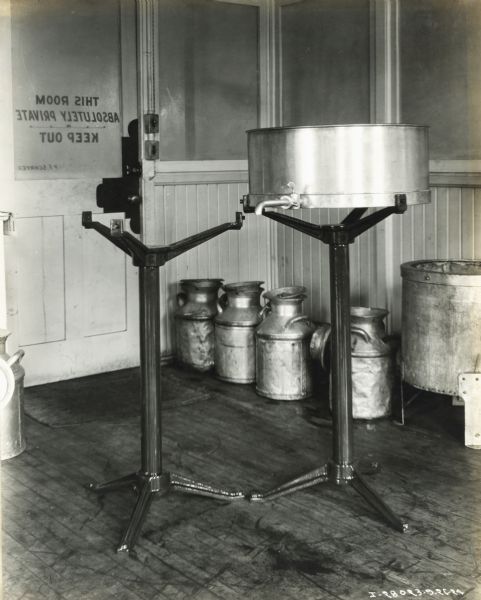 View of two stands in a room, one holding a metal container with a spout. Milk pails are sitting along the wall in the background, and the text on the room's door reads, (backwards): "This Room Absolutely Private, Keep Out; P.F. Schryer."