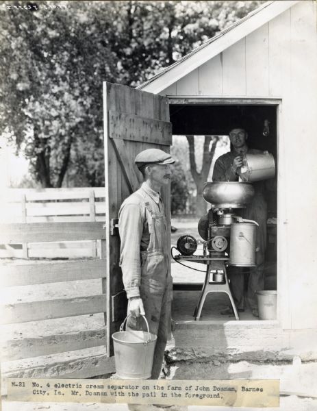 A man is emptying a pail into a Number 4 electric cream separator in a shed on the farm of John Doanan. Mr. Doanan is standing with a pail in the foreground.