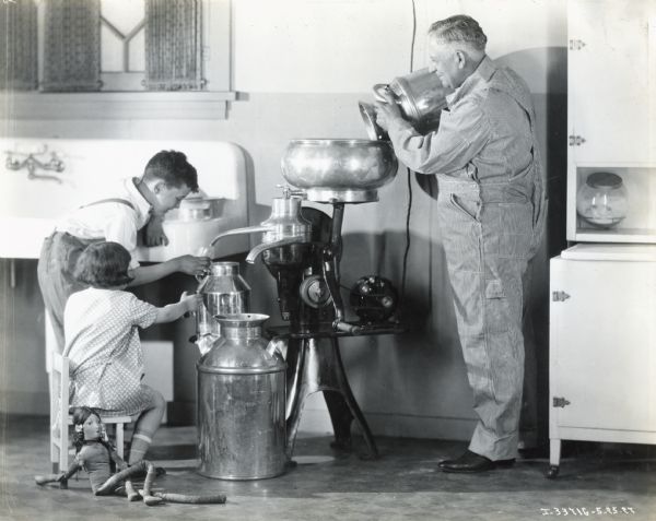 A boy is holding a drinking glass up to the spout of a cream separator as a man is pouring a metal milk pail into the device. A young girl is sitting on a small chair near the boy with her hand outstretched. A doll is lying on the floor at her feet. They are in a large room with a sink and a cabinet.