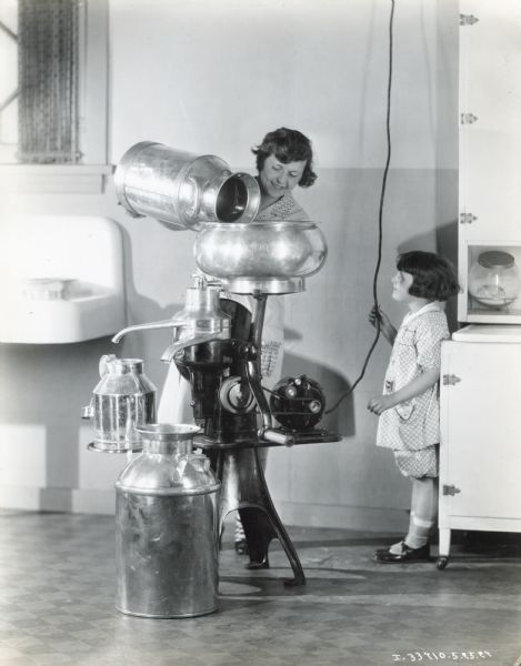 A woman is emptying a metal milk can into a cream separator while a girl is standing by watching. They are in a large room with a sink and a cabinet. An electrical cord is running from the ceiling to the separator's motor.