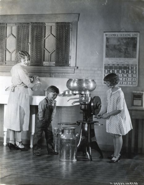 A girl us turning the crank on a cream separator as a boy and a woman are looking on. The woman and children are standing in a kitchen, and the woman is washing a pot at a large sink. A McCormick-Deering calendar is hanging on the wall behind the children.