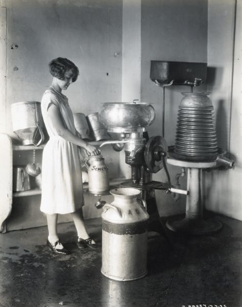 A young woman wearing a dress and a pearl necklace fills a metal pail marked "School" from a cream separator. Additional milk pails are on the floor beside the cream separator, and also lining a shelf in the background.