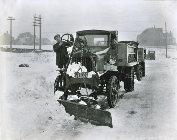Two men using an International truck outfitted with a scraper to remove snow from a road. One man is sitting in the cab to steer the truck, the other man is standing on a running board on the passenger side and steering the plow with a steering wheel assembly that is mounted on the truck hood.