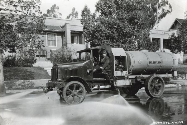 A man sits behind the wheel of an International Model 94 truck owned by the City of Los Angeles. It is marked with the text: "Eng. Dept. 384."  The truck is spraying water for street cleaning in a residential neighborhood.