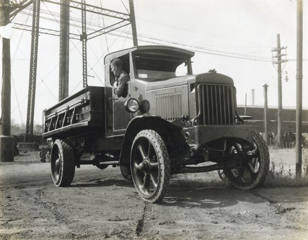 A man looks out the passenger-side window towards the back of an International heavy duty truck (possibly a model 103). There is a ladder attached to the side of the vehicle. In the background is the base of an industrial tower.