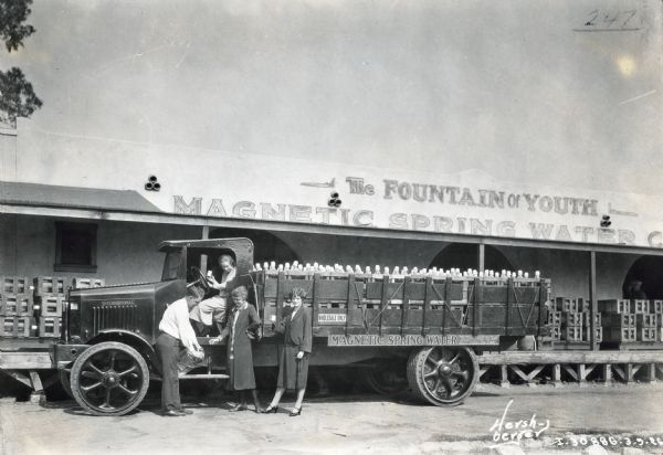 A man stands next to two women near the driver's side door of an International Model 94 truck. Another woman is sitting behind the wheel.  The truck is parked in front of the Magnetic Spring Water Company building and the sign above the building reads: "The Fountain of Youth."
