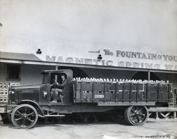 A man sits behind the wheel of an International Model 94 truck loaded with bottles of water. The truck is parked in front of the Magnetic Spring Water Company building, and a sign above the building reads: "The Fountain of Youth."