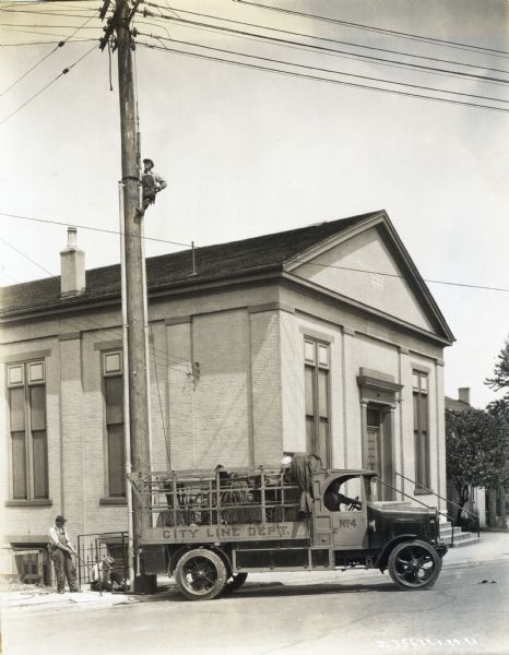 A man climbs a telephone pole next to a building while two others work near its base. Another man sits behind the wheel of an International truck marked: "City Line Dept. No.4" which is backed up next to the pole.