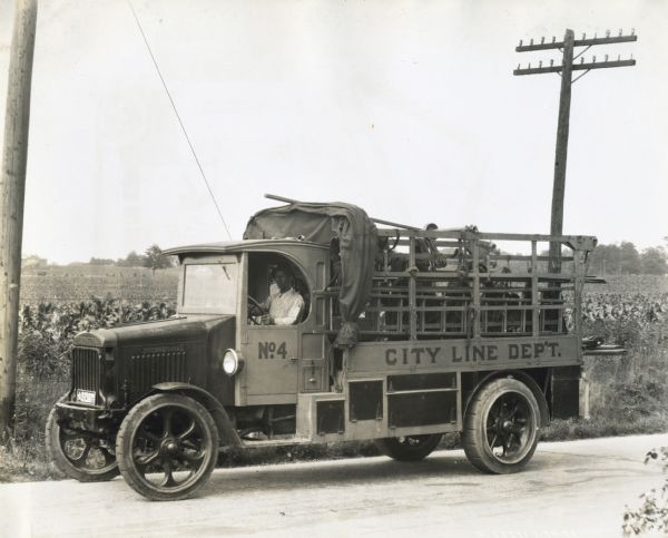 A man sits behind the wheel of an International truck marked: "City Line Dep't. No.4." The truck is parked on a rural road and a telephone pole is in the background.