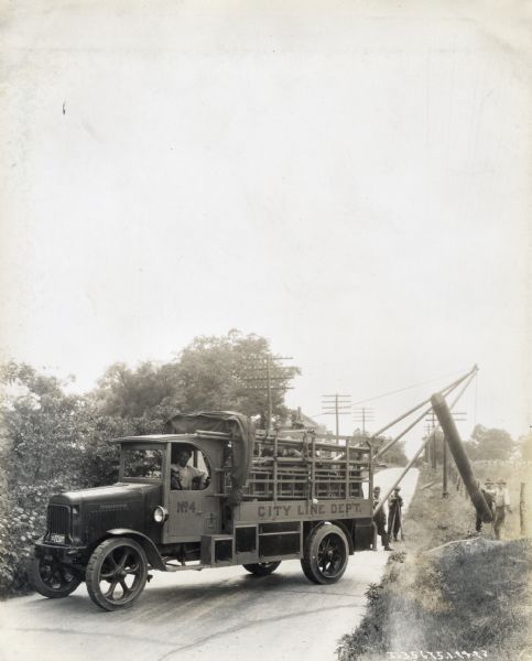 A man sits behind the wheel of an International truck marked "City Line Dept. No.4" Other men raise what appears to be a telephone pole along the side of a rural road with the help of a boom attached to the back of the truck.