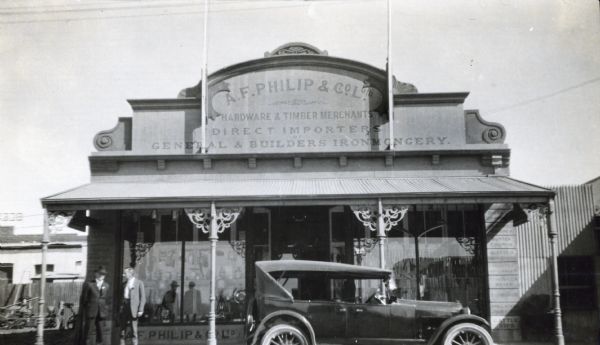View from street of two men standing near the A.F. Philip & Co. Ltd. storefront. The company was likely an International Harvester dealership. An automobile is parked directly in front of the building entrance. The photograph was taken in South Africa.