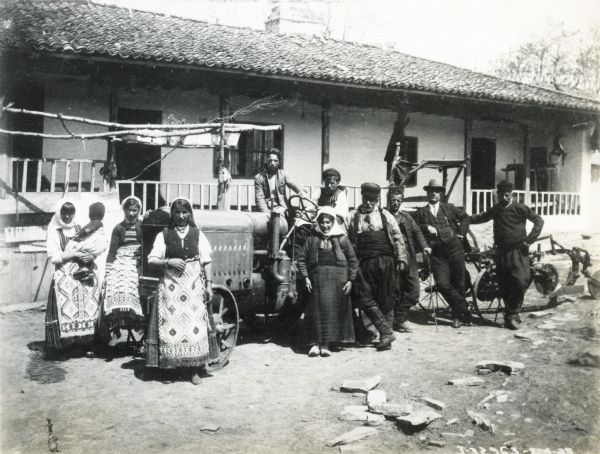 A group of men and women in traditional dress pose outdoors around a Deering tractor and other farm equipment in front of a residence. The scene may be in a foreign country.