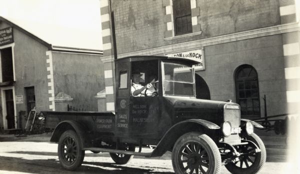 A man drives an International Red Baby truck down a commercial street in South Africa. There is a sign for Nelson and De Kock, an International Harvester (Deering) dealership, on the front of the building behind the truck.