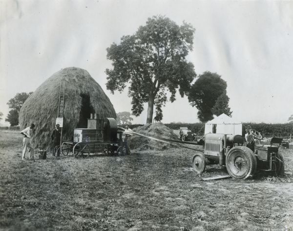Men use an International tractor to power a belt for work with hay. A ladder leans against a large haystack in the background with a sign reading: "Crop Drying Demonstration, Ministry of Agriculture."