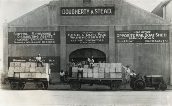 Men sit on boxes loaded onto trailers pulled by an International industrial tractor. The vehicle is parked in front of what appears to be a warehouse building marked "Dougherty & Stead, Shipping Forwarding and Distributing Agents . . . Bond and Duty Paid Warehousement."