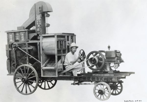 A man wearing a hat is sitting behind the wheel of a tractor or thresher[?] powered by a McCormick-Deering engine. The machine has East Asian lettering on it.