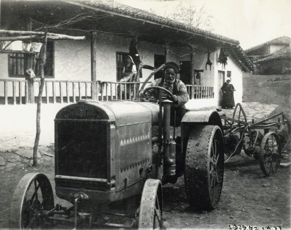 A man drives a Deering tractor, while two women stand near a residential building in the background. The man and women wear what appears to be traditional dress, possibly in a foreign country.
