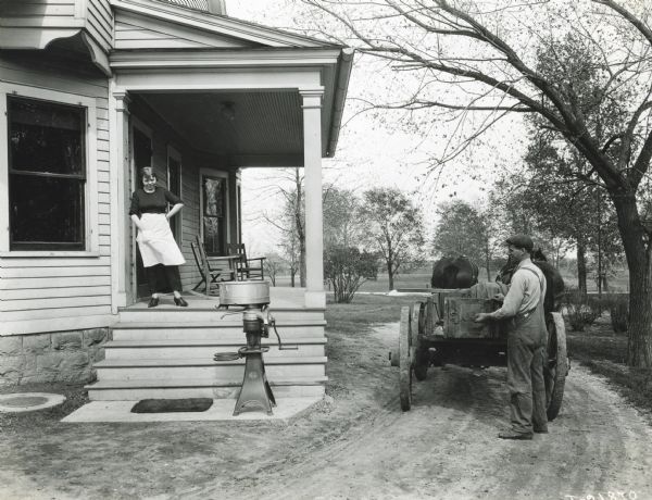 A woman wearing an apron is standing on the porch of a farmhouse to look at a McCormick cream separator at the base of the stairs. Nearby a man wearing overalls is standing at the back of a horse-drawn wagon in the driveway holding onto the wagon gate.