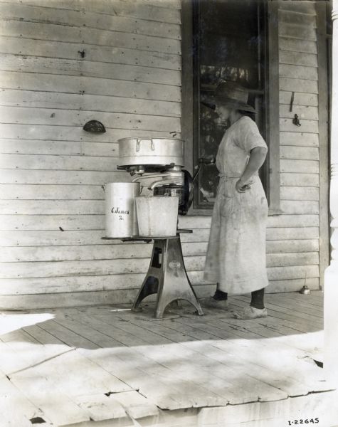 A woman wearing a wide-brimmed hat and a dress is standing and holding the hand crank of a McCormick Primrose cream separator on the porch of a house.