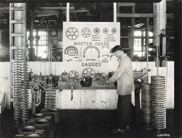 A man is standing at a work table in a factory setting surrounded by metal gears and other parts, possibly for cream separators. A display board attached to a wire barrier features text that reads: "Master Gears" and "Gauges."