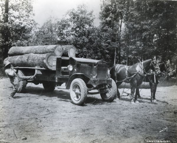 A man loads logs onto the bed of an International truck in a wooded area while two horses stand beside the vehicle.