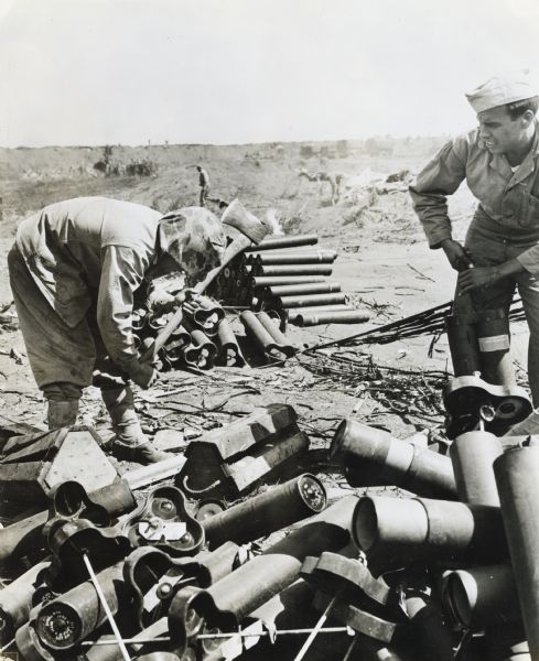 Private First Class Anthony R. Buzzelli opens an ammunition box with an axe for Marines battling on Iwo Jima. Another man on the right appears to be opening a container.