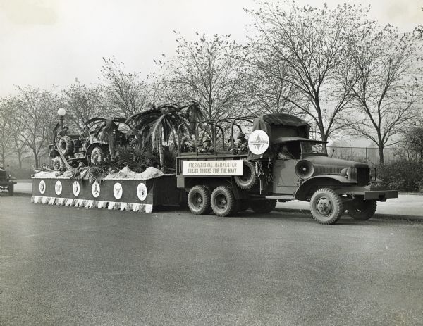 Uniformed naval soldiers sit in an International truck pulling a parade float with a sign reading "International Harvester Builds Trucks for the Navy." The float is decorated with a truck and palm trees.
