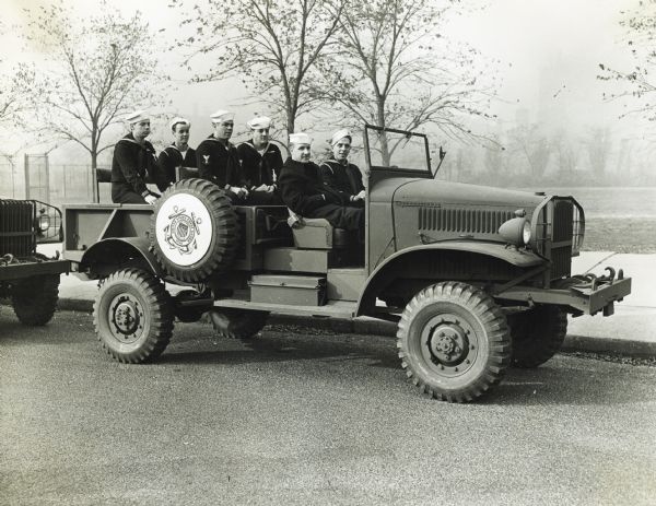 Men from the Coast Guard sit in a military truck parked on the side of a road as part of a parade. The truck was likely made by International Harvester.