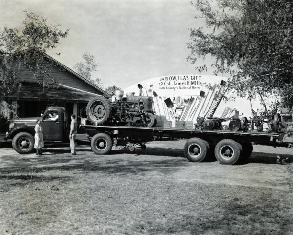 A man and a woman standing in a driveway speak with the driver of an International truck loaded with a tractor, farm implements, and a descriptive display. The tractor and farm implements were presented to Corporal James H. Mills in recognition of his war service. The items were purchased by the citizens of his home town, Bartow, Florida. The sign on the float reads: "Bartow, FLA's Gift To Cpl. James H. Mills, Polk Country's National Hero."