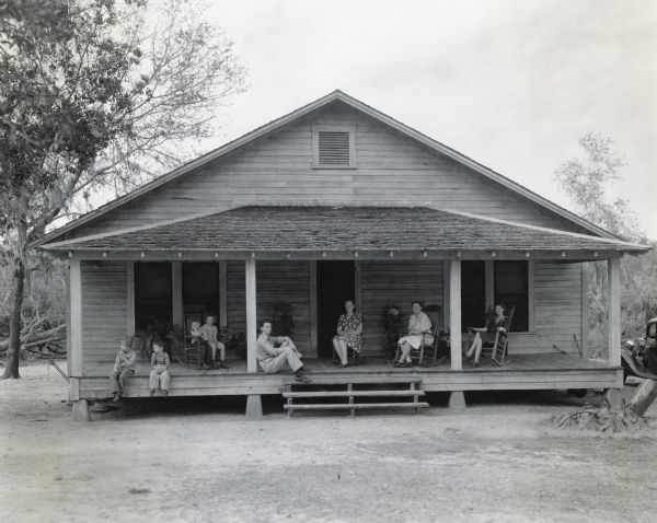 Men, women, and children sit on rocking chairs or on the wooden floor of the front porch of a house. A truck is parked to the right of the home. The home is likely that of Corporal James H. Mills, a war hero honored by his home town of Bartow, Florida with a gift of a Farmall tractor and farm implements.
