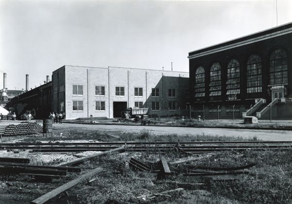 Exterior view of the annex to International Harvester's Ordnance Steel Foundry with railroad tracks running through the foreground. Several men stand near the building and a truck is parked nearby.
