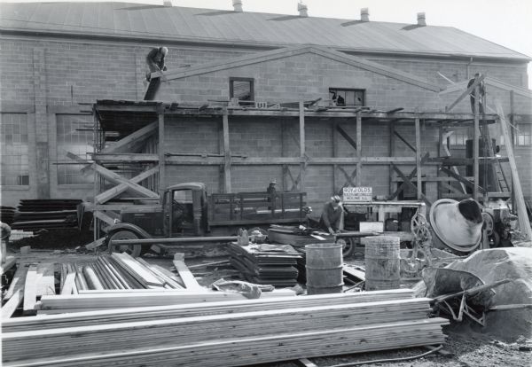 Men are standin on scaffolding to work on the Boiler House at International Harvester's Quad Cities Tank Arsenal. Another man is working on the ground beside a truck and piles of lumber. There is also construction equipment on the grounds.