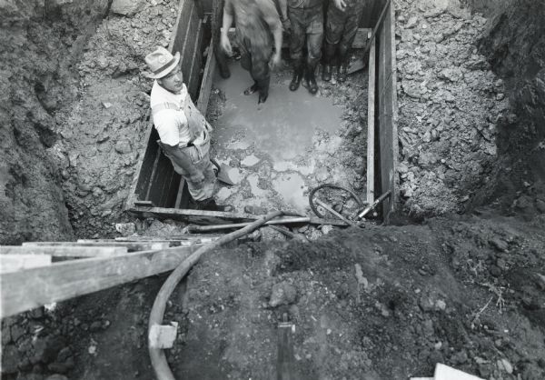 Overhead view of four people standing in a muddy excavated hole for a footing at the Ordnance Steel Foundry. The Foundry may have served as part of International Harvester's war production.