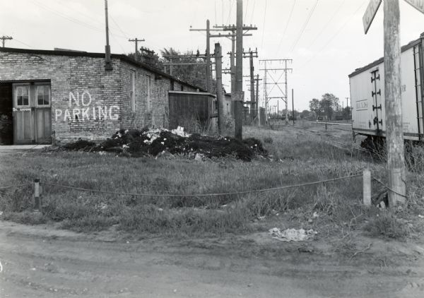 View looking East toward the proposed south boundary fence line of International Harvester's Quad-Cities Tank Arsenal. A railroad track and car are on the right. The brick building on the left has "No Parking" painted on its side.