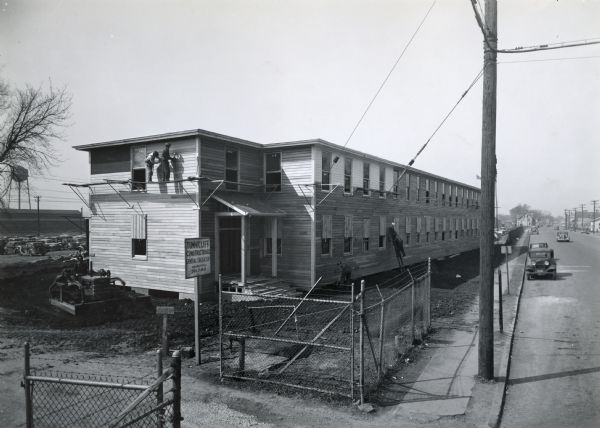 Workers from the Tunnicliff Construction Company build a temporary office building at International Harvester's Bettendorf Tank Arsenal. On the right a parking lot and buildings are along a road that is stretching to the horizon.