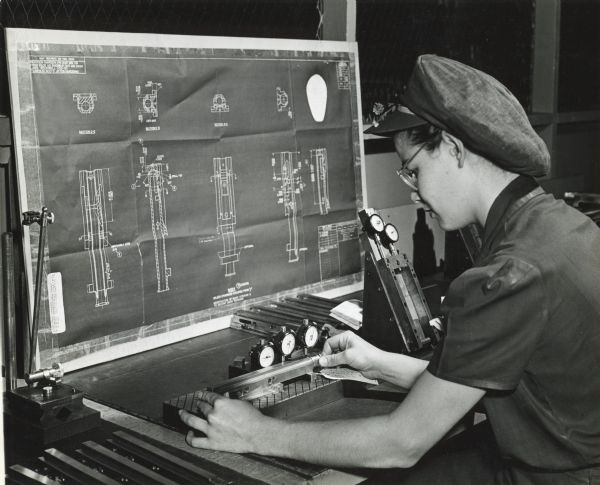 Factory worker Joan Harris sits at a workspace to inspect a 20 mm cannon bolt body part at International Harvester's St. Paul Works. Tools are on the table and a blueprint is on a board in front of her.
