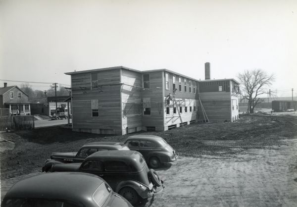 Exterior view of the east side of a temporary office building under construction at International Harvester's Bettendorf Tank Arsenal. Several cars are parked in the dirt lot in the foreground. A man sits on scaffolding to work on the building as another man leans out a window.