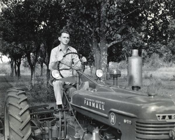 A man, possibly Corporal James H. Mills, operating a Farmall H tractor in what appears to be an apple orchard. Corporal Mills was presented with a tractor by his hometown in recognition of his war service.