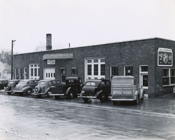 Automobiles and trucks are parked in a row in front of a former International Harvester dealership. A sign on the side of the building advertises 7 up soda.