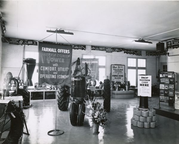 Dealership show room filled with farm equipment and a Farmall M tractor at F.G. Taylor Farm Equipment Co. A banner over a window reads: "FARMALL OFFERS SMOOTH 4 CYLINDER POWER with COMFORT, UTILITY and OPERATING ECONOMY." A display of binder twine includes a sign that reads "Drop Registration Cards Here."
