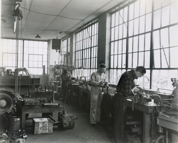 Three men working in a service shop of an International Harvester dealership. The men are standing at work tables with vises along a row of large windows.