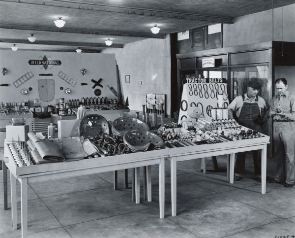 Men stand near an exhibit of tractor belts, seats, supplies and other parts in the showroom of an International Harvester dealership.