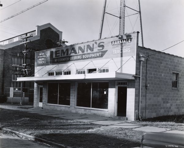 Exterior view of storefront of Lemann's McCormick-Deering Equipment, an International Harvester dealership operated by B. Lemann & Bro., Inc. A large industrial building is on the left.