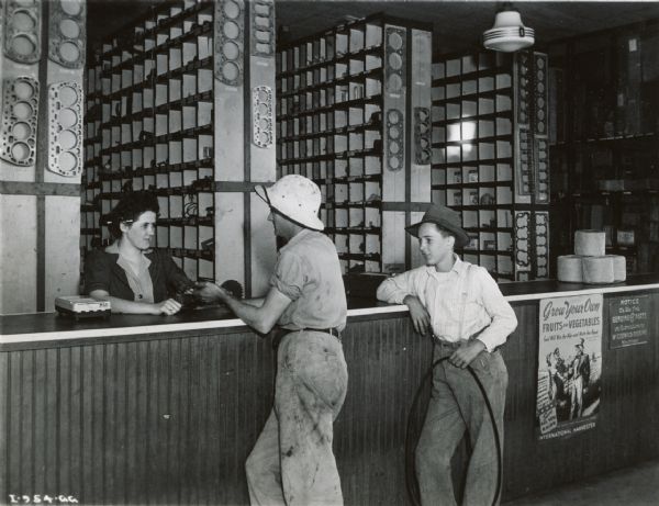 A man and a boy standing at a parts counter in an International Harvester dealership. A woman is helping them from behind a counter. A poster advertising victory gardens, titled: "Grow Your Own Vegetables" is on the front of the parts desk, under a small binder twine display.