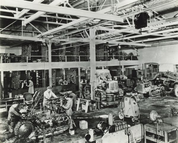 Elevated view of men working on crawler tractors (TracTracTors) and construction equipment in the service shop of J.D. Adams Co., an International Harvester "industrial power" dealership. In the background men work near shelves of parts which are stored on two levels.